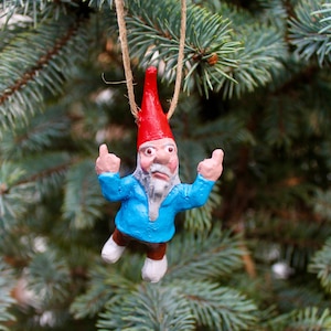 May include: A small, blue gnome ornament with a red hat and a white beard. The gnome is giving the middle finger with both hands. The ornament is hanging from a tree branch.