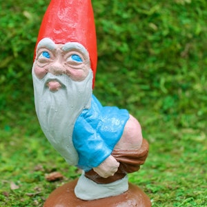 Può includere: Un gnomo da giardino con un cappello rosso, una barba bianca e una camicia blu è seduto su una base marrone. Il gnomo è raffigurato in posizione accovacciata, con le mani che tengono i suoi pantaloni.