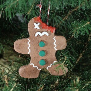 May include: A brown gingerbread man Christmas ornament with white icing and green buttons. The gingerbread man is missing a bite and has red paint on its face, as if it has been injured.