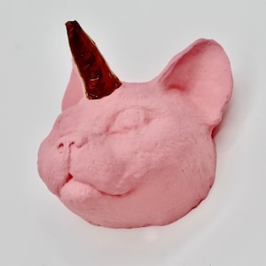 May include: A pink ceramic sculpture of a cat's head with a gold and red horn. The sculpture is a whimsical and modern take on a traditional animal figurine.
