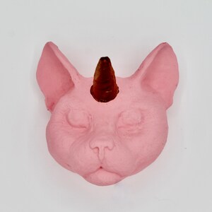 May include: A pink ceramic cat head with a brown horn on top. The cat has closed eyes and a small nose.