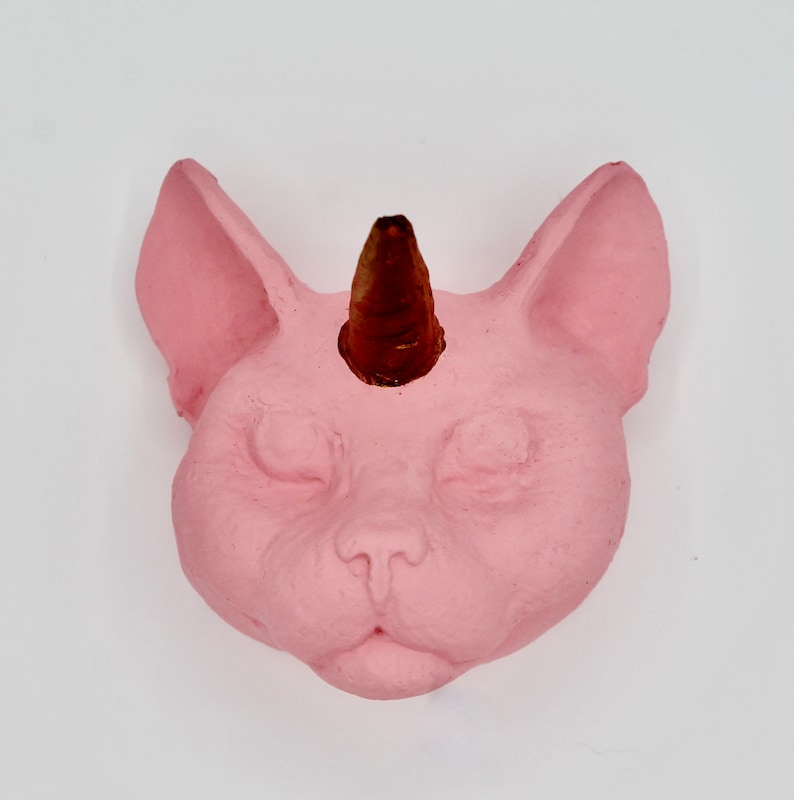 May include: A pink ceramic cat head with a brown horn on top. The cat has closed eyes and a small nose.