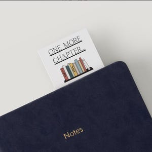 Op de afbeelding: Een marineblauw notitieboek met het woord "Notes" in gouden letters. Een witte bladwijzer met de tekst "ONE MORE CHAPTER..." en een afbeelding van een boekenplank is in het notitieboek geplaatst.
