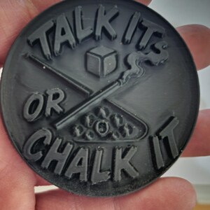 May include: A black, circular object with the words "TALK IT" and "OR CHALK IT" in raised lettering. The design includes a cube, crossed pool cues, and a rack of billiard balls.