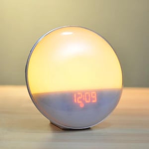 Puede incluir: Un despertador de amanecer blanco y naranja con una pantalla digital que muestra la hora 12:09. El reloj tiene forma redonda con una luz naranja brillante.