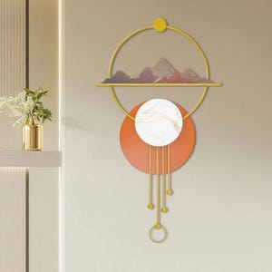 Puede incluir: Arte mural abstracto con un marco circular amarillo y una silueta de montañas. Un círculo naranja sostiene un círculo blanco con efecto mármol. Líneas y esferas doradas cuelgan del círculo naranja, creando un diseño único.