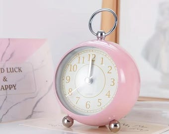Puede incluir: Un reloj despertador analógico rosa, esférico, con asa y patas plateadas. La esfera del reloj es blanca con números y manecillas negras. El reloj está sobre una superficie blanca.