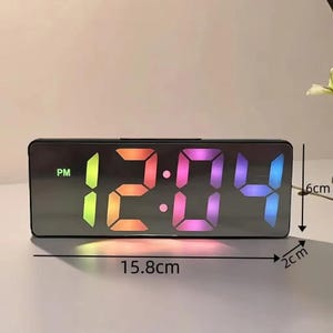 May include: A rectangular digital alarm clock displaying "12:04 PM" in a rainbow gradient. The clock is black, with dimensions of 15.8 cm long, 6 cm high, and 2 cm wide.