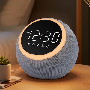 May include: A gray fabric-covered digital alarm clock displaying 12:30 AM. The clock has a circular display with a glowing orange border and several icons. It sits on a wooden surface next to a small plant.