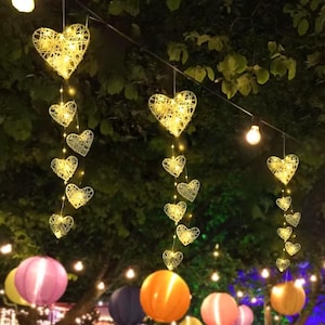 May include: Decorative string lights featuring woven heart-shaped lanterns illuminated with warm white lights. The hearts are suspended from a wire, creating a festive and romantic ambiance. Colorful paper lanterns are visible in the background.