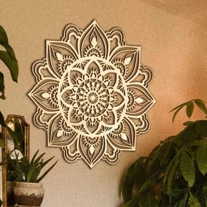 Puede incluir: Una pieza de arte mural de mandala de madera con diseños intrincados y en capas. Los tonos de madera natural crean una estética cálida y neutra. El mandala está montado en una pared de color neutro.