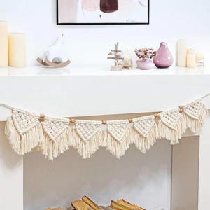 May include: Cream-colored macrame banner with triangular shapes and fringe. The banner is strung with wooden beads and hangs against a white background. Decorative home decor.