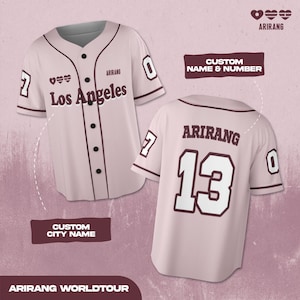 Op de afbeelding: Een lichtroze honkbalshirt met de tekst "Los Angeles" en het nummer "0" op de voorkant. De achterkant van het shirt toont "ARIRANG" en het nummer "13". Het shirt heeft donkerbruine accenten en de tekst "CUSTOM NAME & NUMBER".
