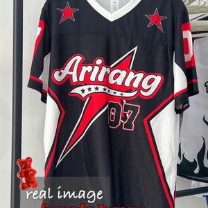 May include: Black jersey with red and white accents, featuring the word "Arirang" in white script with red accents. The jersey has red stars and the number "07". The text "real image Sample Jersey" is at the bottom.