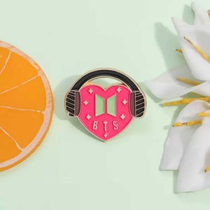 May include: A pink heart-shaped pin with the letters "BTS" at the bottom, framed by black headphones. The heart features two green rectangles and small diamond accents. The pin is set against a light green background, with a slice of orange and white flowers.