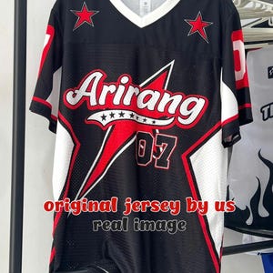 Peut inclure: Un maillot noir à col en V avec des accents blancs et rouges, un grand motif d'étoile rouge, le texte Arirang et le numéro 07. Le maillot porte l'inscription original jersey by us real image. Le tissu présente une texture en maille filet.