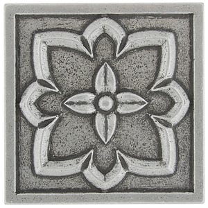 May include: Square, silver decorative tile with a raised floral design. The design features a central four-petal flower surrounded by stylized leaves and a textured background. The tile has a square border.