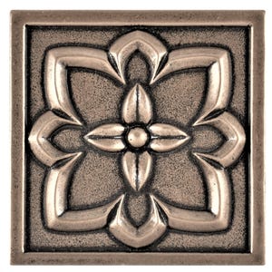 May include: Square, bronze-coloured decorative tile with a raised floral design. The central flower has four petals surrounding a small, circular centre. The corners of the tile are also square, and the overall texture is slightly rough.
