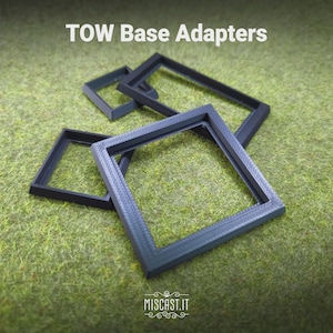 Puede incluir: Tres adaptadores de base cuadrados y rectangulares de plástico negro con la etiqueta TOW Base Adapters. Estos componentes presentan un acabado de superficie texturizado y están diseñados con un centro hueco, apilados sobre una superficie verde.