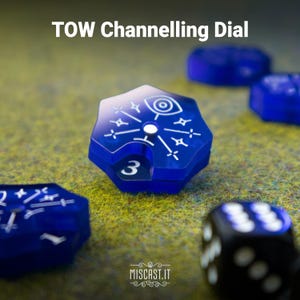 May include: A blue translucent hexagonal TOW Channelling Dial featuring a white eye and star graphic on the top surface with a number 3 visible in the side cutout. The dial is positioned among other blue hexagonal game components and a black die.