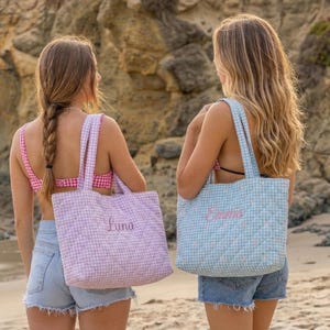 May include: Two quilted tote bags with gingham patterns. One is light purple, the other is light blue. Each bag has a name embroidered on it in a cursive font. The bags have matching straps and are carried on the beach.