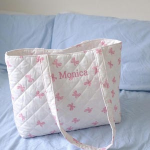 Puede incluir: Bolso tote acolchado blanco con estampado de mariposas rosas y el nombre "Monica" impreso en rosa. El bolso tiene un patrón de diamantes y asas a juego. El bolso está sobre una superficie azul claro.