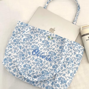 May include: A floral print tote bag in white and light blue, with matching handles. The bag contains a laptop and a white and beige insulated water bottle. The name "Olivia" is embroidered on the front of the bag.