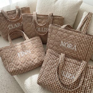 May include: Five quilted tote bags in a brown and white gingham pattern. Each bag features two handles and is embroidered with a unique name in white capital letters: CLARA, OLLIE, FINN, NORA, and ELIZA. The bags have a soft, padded texture.