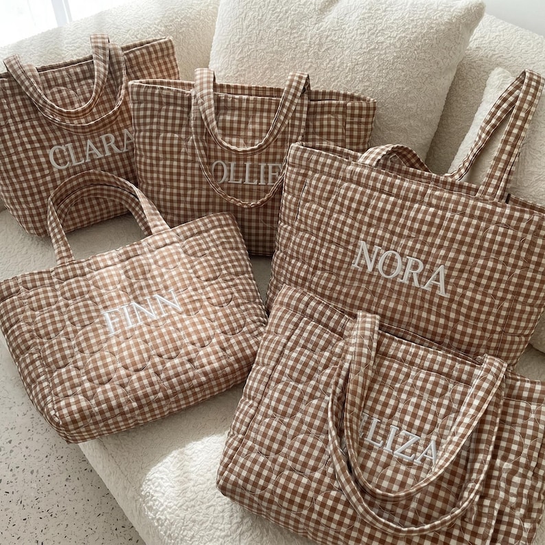 May include: Five quilted tote bags in a brown and white gingham pattern. Each bag features two handles and is embroidered with a unique name in white capital letters: CLARA, OLLIE, FINN, NORA, and ELIZA. The bags have a soft, padded texture.