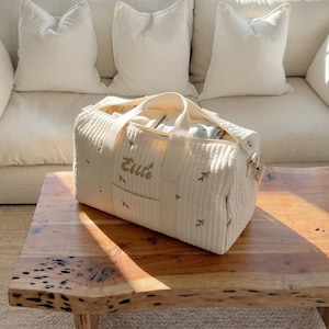 May include: Cream-colored quilted duffel bag with embroidered name "Ellie" and small leaf accents. The bag has a top handle and a front pocket. The bag is filled with items and sits on a wooden table.