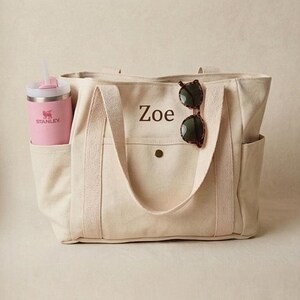 May include: A cream-colored canvas tote bag with the name "Zoe" printed on the front. The bag has a pocket, side pockets, and a pink Stanley water bottle with a straw in one pocket. Tortoiseshell sunglasses rest on the handle.