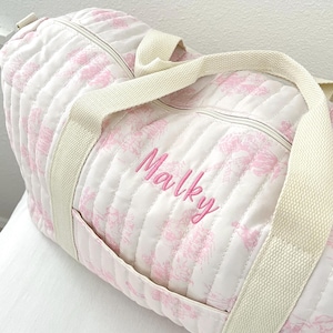 May include: A quilted duffel bag with a white background and a pink floral pattern. The bag has cream-colored handles and a zipper closure. The name "Malky" is embroidered in pink on the side of the bag.