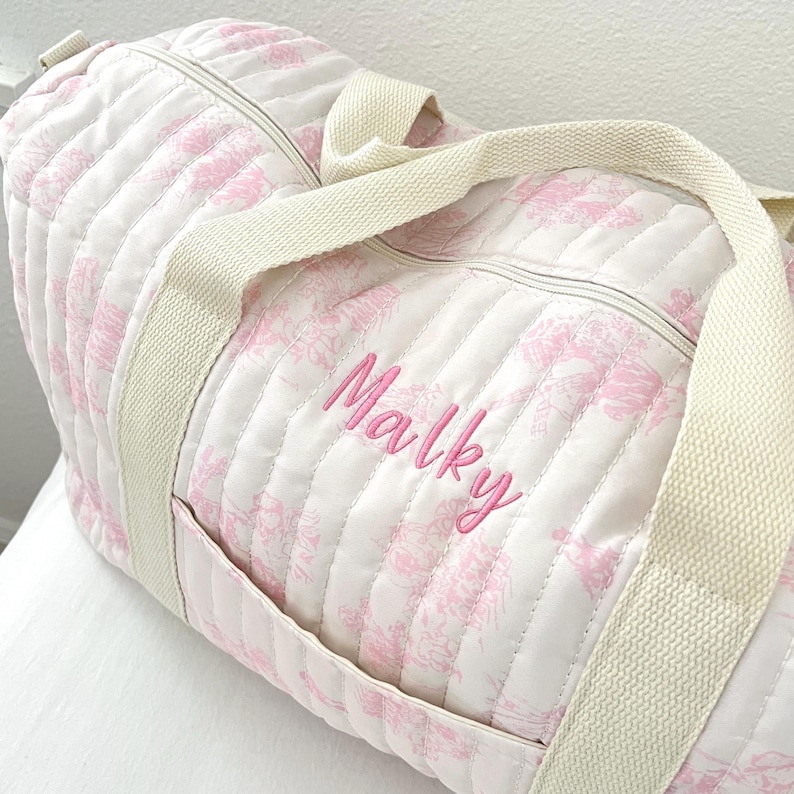 May include: A quilted duffel bag with a white background and a pink floral pattern. The bag has cream-colored handles and a zipper closure. The name "Malky" is embroidered in pink on the side of the bag.