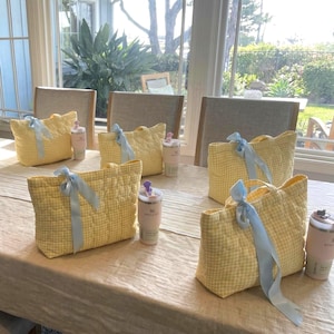 May include: Four quilted tote bags in a yellow and white gingham pattern, each featuring a light blue ribbon tied in a bow on the front handle. Beside each bag sits a cylindrical insulated tumbler in a soft pink or white color with a lid.