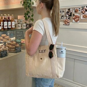 May include: A cream-colored canvas tote bag with the name "LILY" printed on the front. The bag has a side pocket holding a white Stanley tumbler and sunglasses. The person is standing in front of a bakery display.