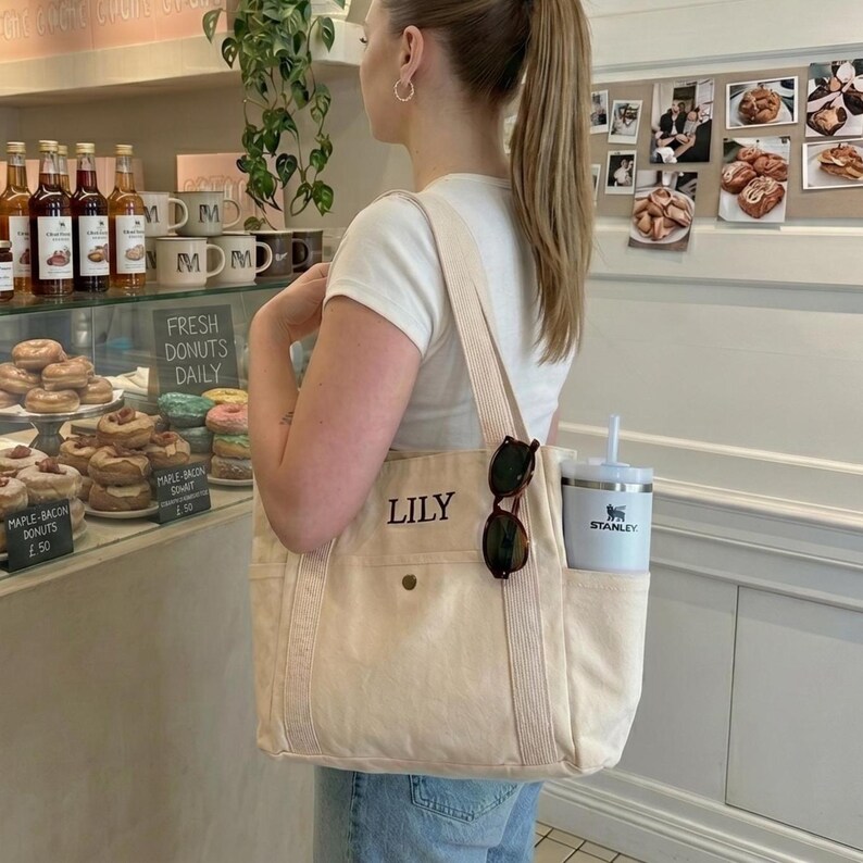 May include: A cream-colored canvas tote bag with the name "LILY" printed on the front. The bag has a side pocket holding a white Stanley tumbler and sunglasses. The person is standing in front of a bakery display.