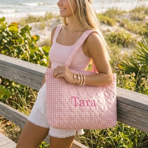 May include: A quilted tote bag featuring a light pink and white gingham pattern. The bag has two shoulder straps and the name Tara embroidered in pink thread on the front. The fabric has a stitched texture throughout the entire surface.