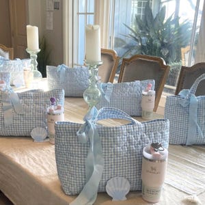 Peut inclure: Plusieurs sacs fourre-tout en vichy bleu clair et blanc avec des poignées en ruban bleu clair sont disposés sur une table. Chaque sac est associé à un gobelet Stanley Quencher rose. De grands bougeoirs en verre contiennent de hautes bougies blanches.