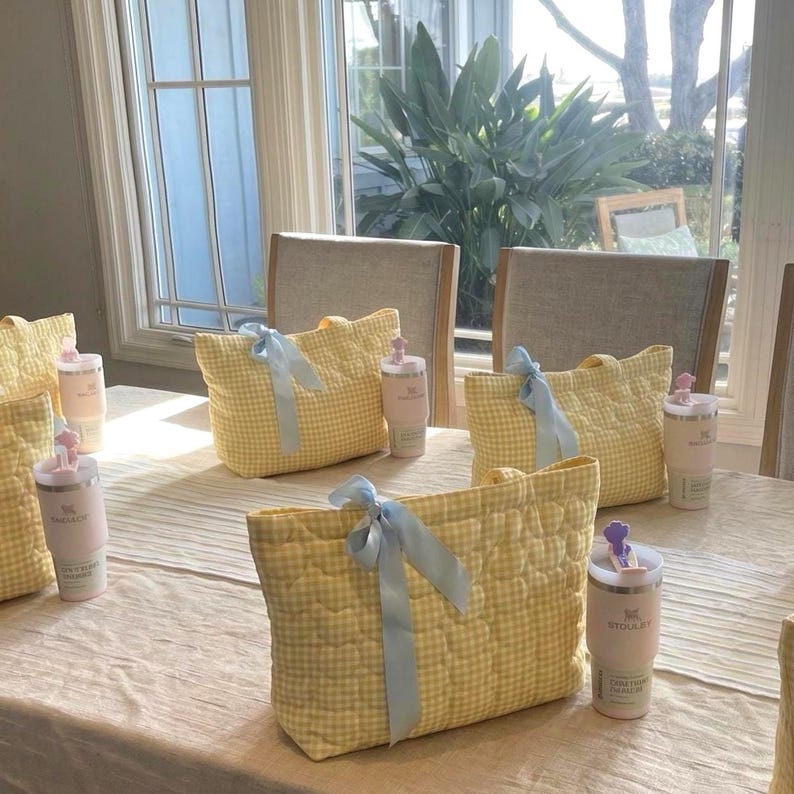 May include: A set of quilted yellow and white gingham tote bags, each adorned with a light blue satin ribbon bow. Accompanying each bag is a pale pink insulated tumbler with a matching lid and a small decorative topper on the straw.