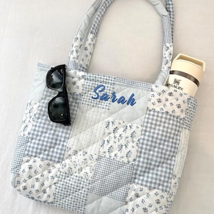 May include: A quilted tote bag with a patchwork design in shades of blue and white. The bag features a floral print, gingham, and solid fabrics. The name "Sarah" is embroidered in blue. Black sunglasses and a Stanley thermos are inside.