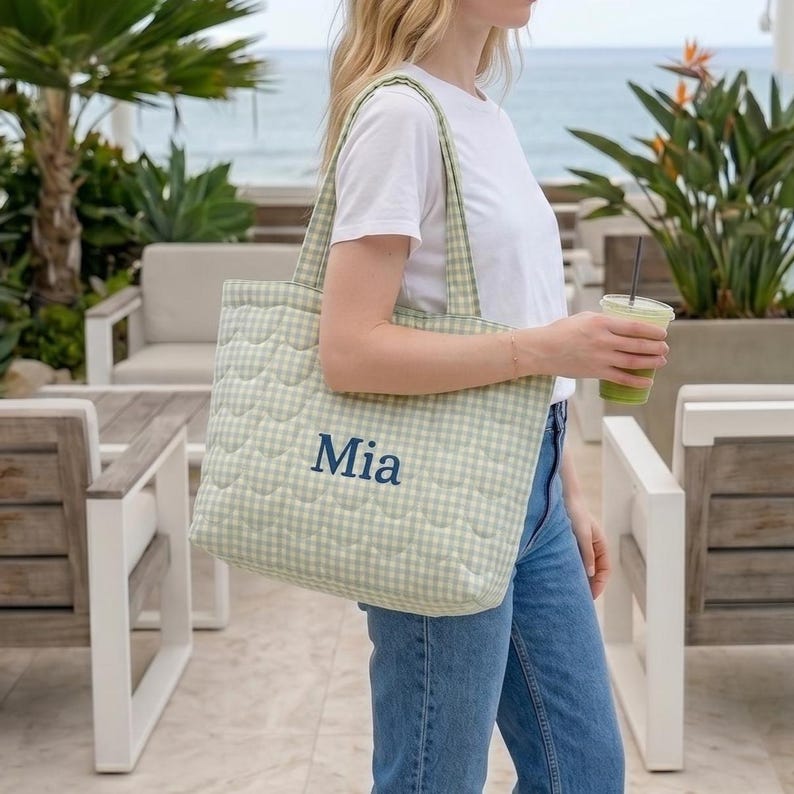 May include: A quilted tote bag featuring a light green and white gingham pattern. The bag has two shoulder straps and the name Mia embroidered in dark blue thread on the front center. The quilted texture consists of a repeating scalloped pattern.