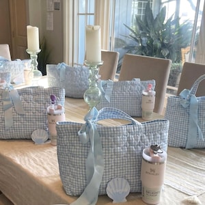 May include: Multiple quilted tote bags featuring a light blue and white gingham pattern, each adorned with a light blue ribbon bow. Accompanying each bag is a light pink insulated tumbler with a straw and a small shell-shaped tag with text.