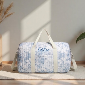 May include: A white and blue floral patterned duffel bag with the name "Ellie" embroidered in blue. The bag has cream-colored handles and a shoulder strap. The bag is on a neutral-colored rug.