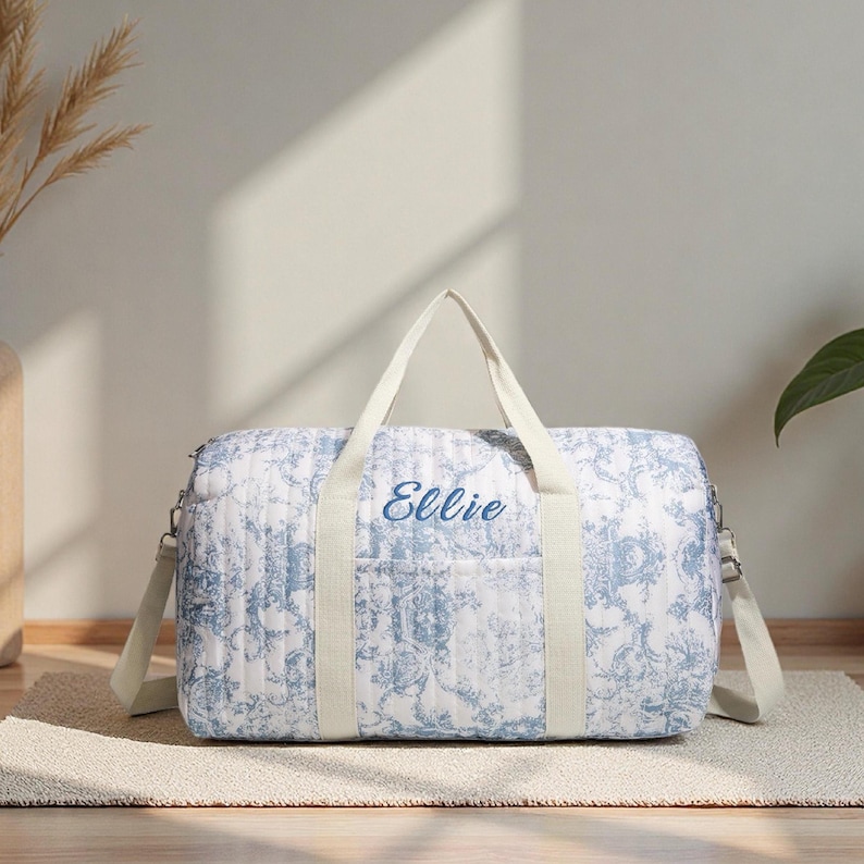 May include: A white and blue floral patterned duffel bag with the name "Ellie" embroidered in blue. The bag has cream-colored handles and a shoulder strap. The bag is on a neutral-colored rug.