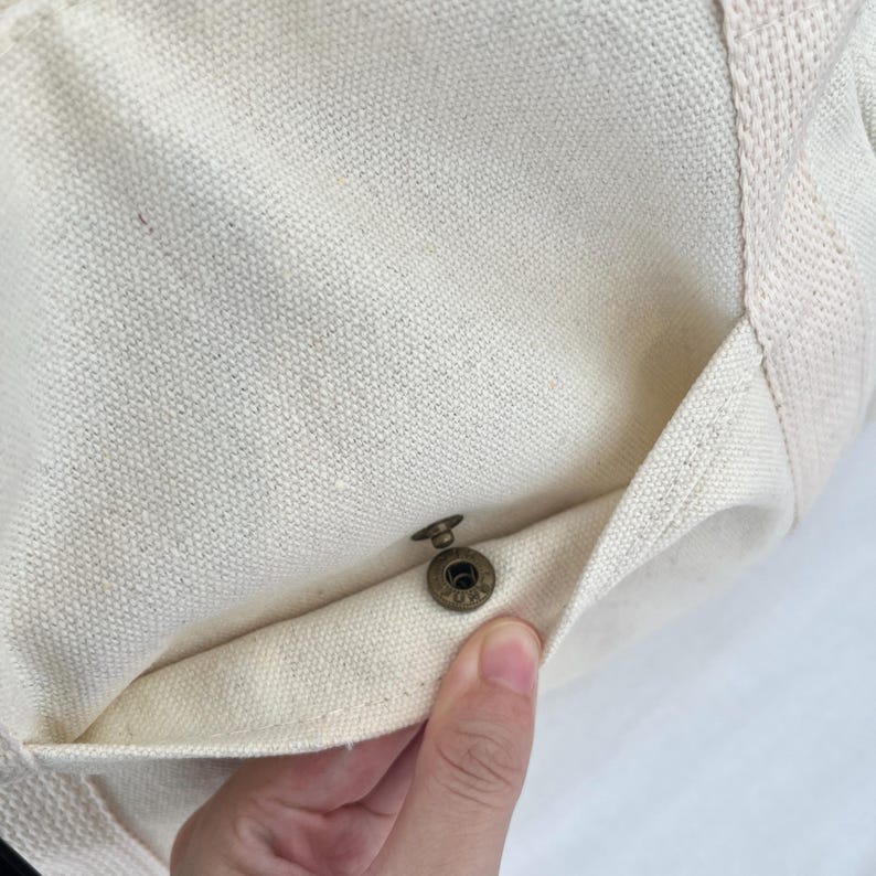 May include: Close-up of a cream-colored canvas tote bag with a small pocket. The pocket is secured with a bronze-colored snap closure. The bag's fabric has a textured appearance.