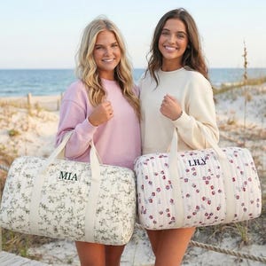 May include: Two women stand on a beach, each holding a personalized duffel bag. One bag has a floral pattern with "MIA" embroidered, the other has a floral pattern with "LILY". The bags have cream-colored handles.
