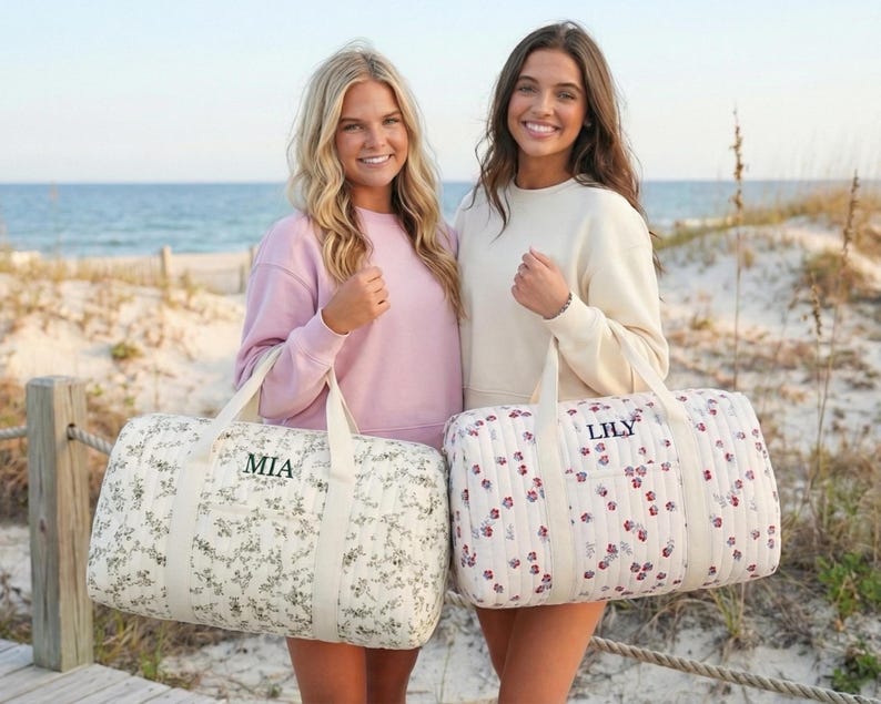 May include: Two women stand on a beach, each holding a personalized duffel bag. One bag has a floral pattern with "MIA" embroidered, the other has a floral pattern with "LILY". The bags have cream-colored handles.