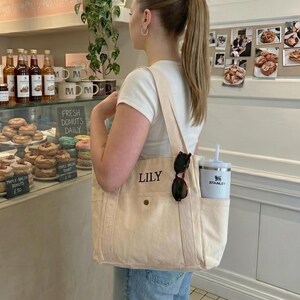 Op de afbeelding: Een cr&egrave;mekleurige draagtas met de naam "LILY" op de voorkant geborduurd. De tas heeft een zijvak met een witte Stanley-beker en zonnebril. De tas wordt gedragen door een persoon in een wit T-shirt en een blauwe jeans.