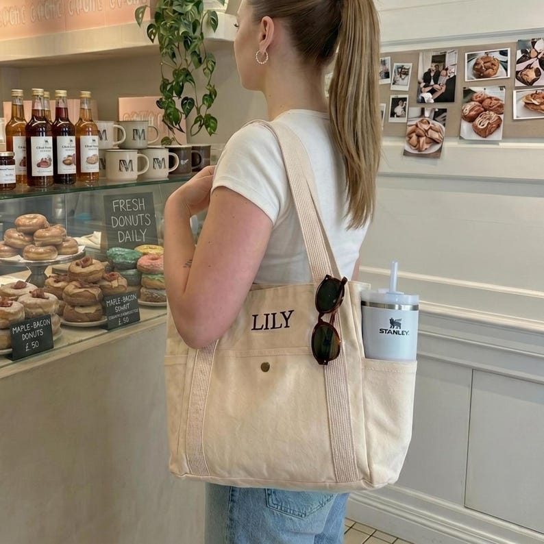 Op de afbeelding: Een cr&egrave;mekleurige draagtas met de naam "LILY" op de voorkant geborduurd. De tas heeft een zijvak met een witte Stanley-beker en zonnebril. De tas wordt gedragen door een persoon in een wit T-shirt en een blauwe jeans.