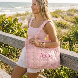 May include: A quilted tote bag featuring a light pink and white gingham pattern with the name Tara embroidered in pink thread on the front. The bag has two matching gingham shoulder straps and a rectangular shape with a soft, padded texture.